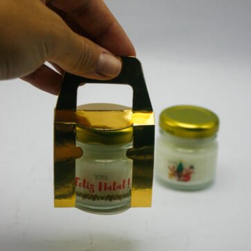 Vela aromática Feliz Natal Candela Aromas sendo segurada, pote de vidro dourado.