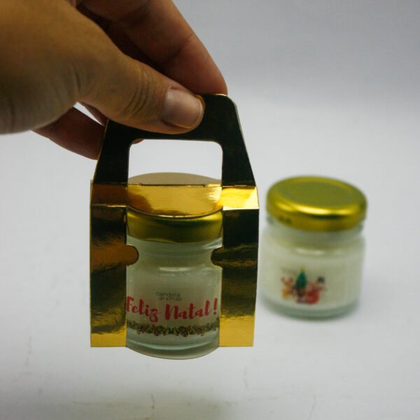 Vela aromática Feliz Natal Candela Aromas sendo segurada, pote de vidro dourado.